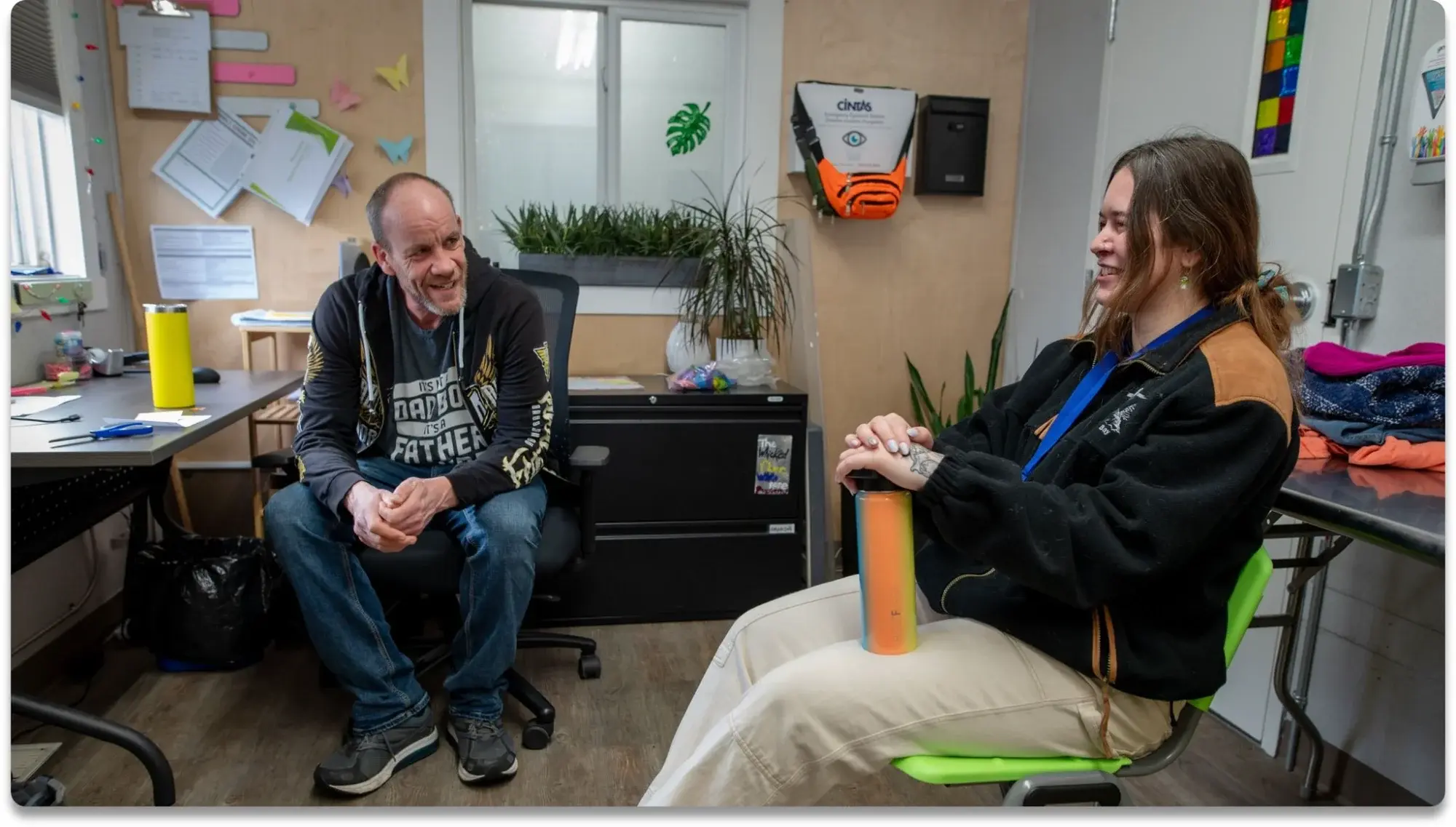 Two people having a friendly conversation in a community office space; an older man with a goatee wearing a graphic t-shirt sits on an office chair while a young woman with long brown hair in a black and tan jacket sits across from him holding a colorful water bottle, surrounded by plants, bulletin boards with papers and origami decorations